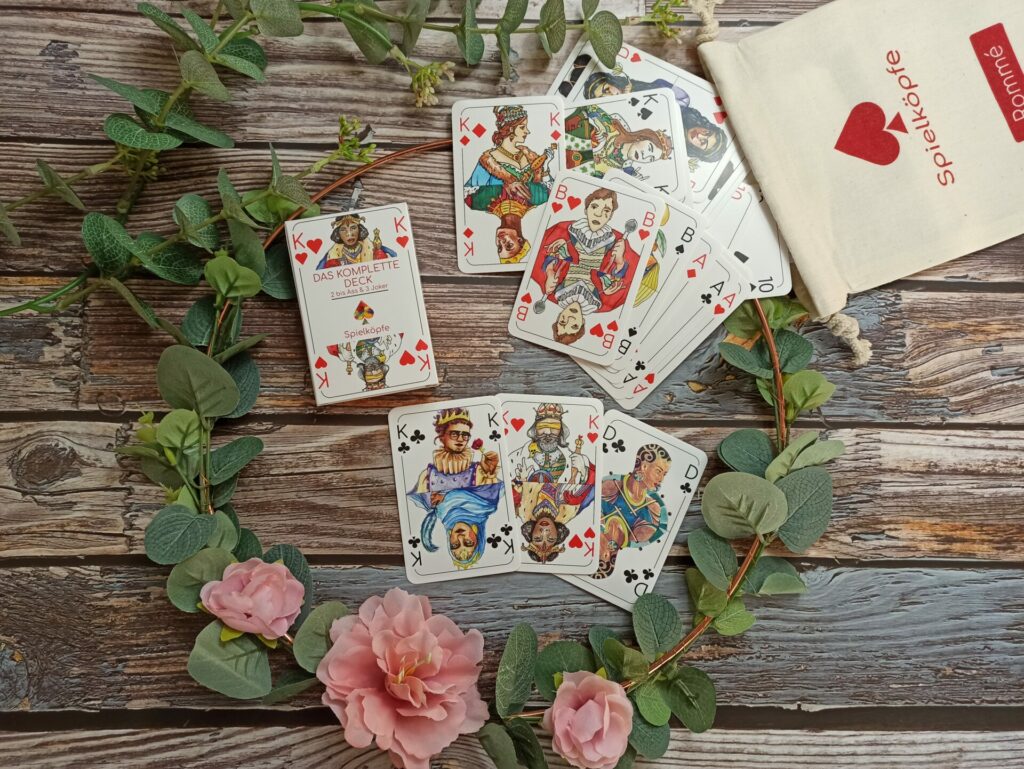 mehrere Spielkarten rutschen aus einem Baumwollbeutel umrahmt von einem Blumenkranz