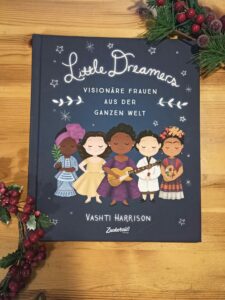 Das Buch "Little Dreamers" ist zu sehen. Es ist dunkelblau, darauf sind 5 niedliche Illustrationen von Frauen zu sehen. Darüber steht in weiß der Titel, sowie der Untertitel "Visionäre Frauen aus der ganzen Welt". Unter der Illustration stehen der Name der Autorin und der Verlag.