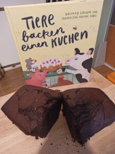 Kinderbuch "Tiere backen einen Kuchen" und davor ein aufgeschnittener Schokokuchen