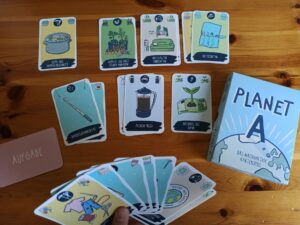 Sieben Spielkarten liegen offen auf dem Tisch, darunter ist verdeckt eine Aufgabenkarte sowie eine Spielerhand mit ebenfalls 7 Karten zu sehen. Rechts liegt die Verpackung des Spiels "Planet A"