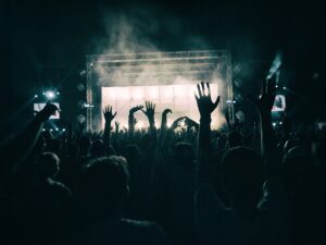 Eine Menschenmenge im dunkeln von hinten. Im Hintergrund in Blickrichtung der Menschen ist eine hell erleuchtete Bühne zu sehen. Die Menschen strecken die Hände in die Luft.