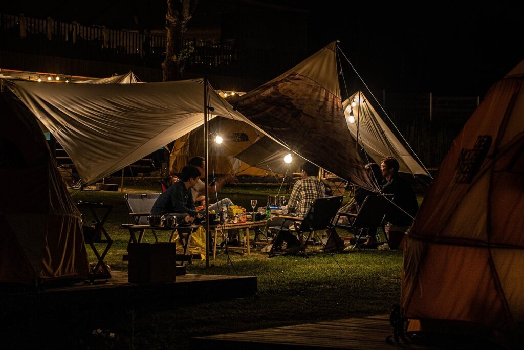 Eine Gruppe Menschen sitzt im Dunkeln unter einer gespannten Zeltplane um einen Tisch und unterhält sich. Zwei Glühbirnen beleuchten die Szene. Außen herum kann man einige Zelte erkennen.